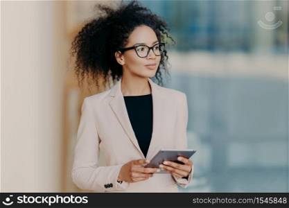 Attractive elegant dark skinned businesswoman uses digital tablet, dressed in formal wear, stands in office, looks thoughtfully aside, reads business news on web page, makes online payments.