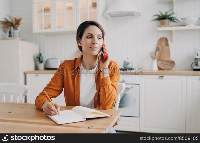 Attractive european woman is entrepreneur. Lady in orange shirt receiving order on phone. Woman is remote worker at home. Girl is sitting at table at kitchen and talking on phone to customer.. Attractive european woman is entrepreneur. Lady in orange shirt receiving order on phone.