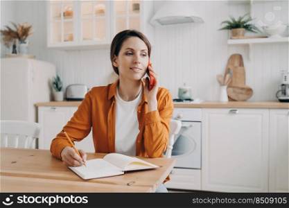 Attractive european woman is entrepreneur. Lady in orange shirt receiving order on phone. Woman is remote worker at home. Girl is sitting at table at kitchen and talking on phone to customer.. Attractive european woman is entrepreneur. Lady in orange shirt receiving order on phone.