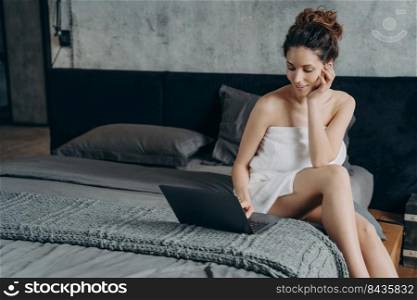 Attractive european woman wrapped in towel after bathing is chatting on pc. Girl takes shower and relaxing in bedroom in her apartment. Remote worker lifestyle. Weekend of happy freelancer.. Attractive happy european woman wrapped in towel after bathing is chatting on pc in bedroom.