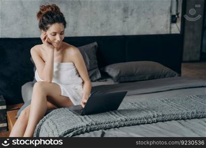 Attractive european woman wrapped in towel after bathing is chatting on pc. Girl takes shower and relaxing in bedroom in her apartment. Remote worker lifestyle. Weekend of happy freelancer.. Attractive happy european woman wrapped in towel after bathing is chatting on pc in bedroom.
