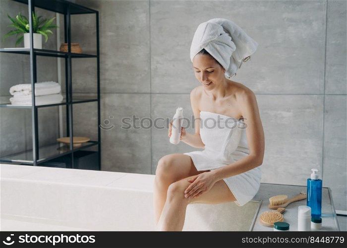 Attractive european woman wrapped in towel applying body lotion to her leg after shower. Young woman is bathing in morning at home. Anti-cellulite massage and bodycare. Enjoyment and relaxation.. Young woman is bathing in morning at home and applying body lotion to her leg after shower.