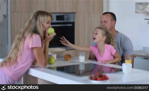 Attractive family with adorable curly blonde daughter enjoying breakfast in kitchen in the morning. Gorgeous mother leaning on table and eating apple while interacting with her handsome husband and little girl in domestic interior.