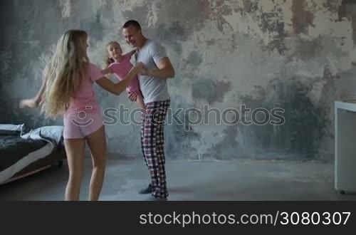 Attractive family with adorable daughter in pajamas having fun at home dancing in the morning. Handsome father carrying cute little girl and holding hands of beautiful wife while dancing together at home in grunge styled room. Stabilized shot. Slo mo