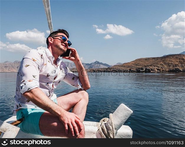 Attractive, fashionable man sitting on a boat and photographing coastal cliffs, blue sky and waves. Concept of leisure and travel. Attractive, fashionable man sitting on a boat