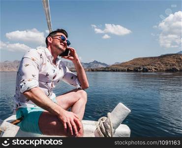 Attractive, fashionable man sitting on a boat and photographing coastal cliffs, blue sky and waves. Concept of leisure and travel. Attractive, fashionable man sitting on a boat