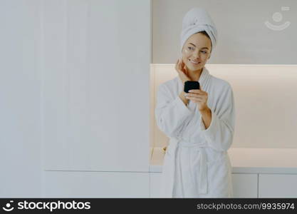 Attractive female applies moisturising cream on face takes care of her complexion and skin holds mirror wears white bathrobe smiles genlty looks thoughtfully poses over modern furniture at home