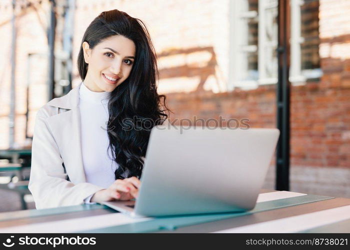 Attractive female blogger typing new challenging post using modern laptop computer while sitting at outdoor cafe. Pretty brunette businesswoman working with laptop doing her business project