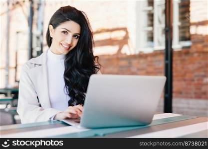 Attractive female blogger typing new challenging post using modern laptop computer while sitting at outdoor cafe. Pretty brunette businesswoman working with laptop doing her business project