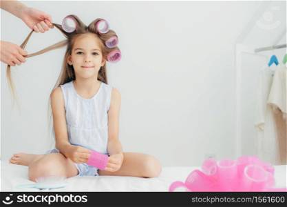 Attractive female child has curlers on head, going to have wonderful hairstyle, recieves beauty lessons from affectionate mother, isolated over white background. Children and beauty concept.