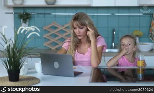 Attractive female entrepreneur in casual clothes working on laptop and smartphone from home office while her cute daughter drinking juice in the kitchen. Busy female freelancer talking with clients on mobile phone in the morning.