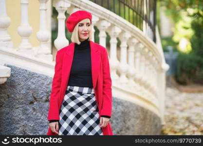 Attractive female in stylish red coat and checkered skirt looking away while standing near white fence on street of city. Fashionable woman near fence in city outdoors