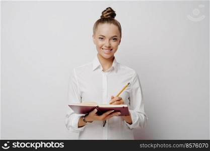 Attractive female journalist with lovely smile in formal clothes listening and writing down notes in red notebook, holding pencil and expressing positive emotions while standing in front of light wall. Business professional in white shirt writing down ideas in note book