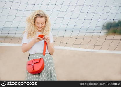 Attractive female model focused into smart phone, reads information on web page, has bright bag, smiles joyfully as recieve mesage from boyfriend, poses on beach. People, lifestyle and rest concept