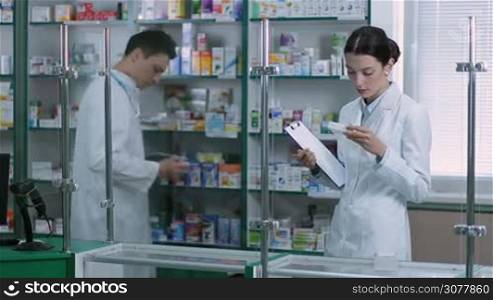 Attractive female pharmacist with clipboard and pencil counting stock with colleague in pharmacy. Female chemist woman checking medicines while her colleague counting medicine stock availability using digital tablet pc on background.