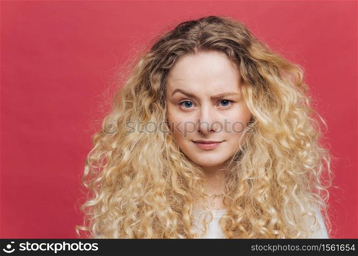 Attractive female with bushy light curly hair, raises her eyebrow, feels puzzlement, can`t undersatnd something, isolated over pink background. Beautiful young surprised woman poses in studio