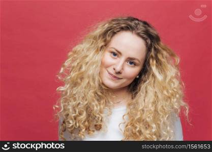 Attractive female with curly bushy hair and blue eyes, looks positively at camera, feels satisfied as meets with friends, isolated over bright pink background. People, beauty and emotions concept