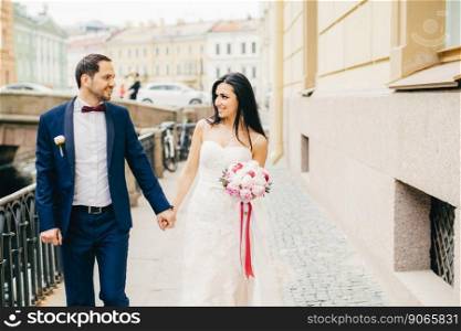 Attractive female with dark hair, wears white dress, holds bouquet, keeps bridegroom s hand, look at each other with great love, have walk across ancient city on bridge. Positive emotions concept