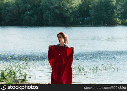 Attractive fit female covering herself with red wrap from unexpected wind blow, mouth wide open, long hair, water beach background. Healthy fun lifestyle, travel, relax, enjoy nature concept