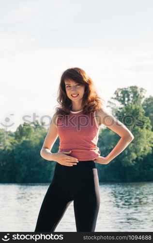Attractive fit female practicing sports activity, yoga, wearing tight clothes, smiling, hand on her belly, long hair, water beach background. Healthy lifestyle, keep fit, weight loss concept