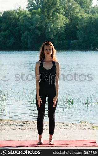 Attractive fit female practicing sports activity, yoga, wearing tight clothes, stretching pose, hands along the body, long hair, outdoor water background. Healthy lifestyle, keep fit, weight loss concept