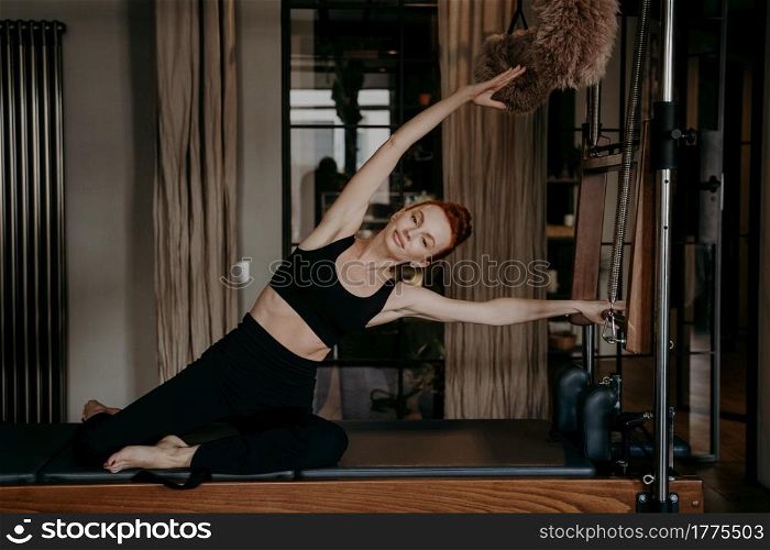 Attractive fit red haired woman with perfect body shape exercising and stretching on pilates cadillac while having fitness training in modern cozy studio. Sport and healthy lifestyle concept. Young athletic woman during pilates training