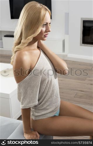 Attractive, fresh, natural woman with blonde, long, wavy hair in beige sweater is sitting on the sofa in living room.