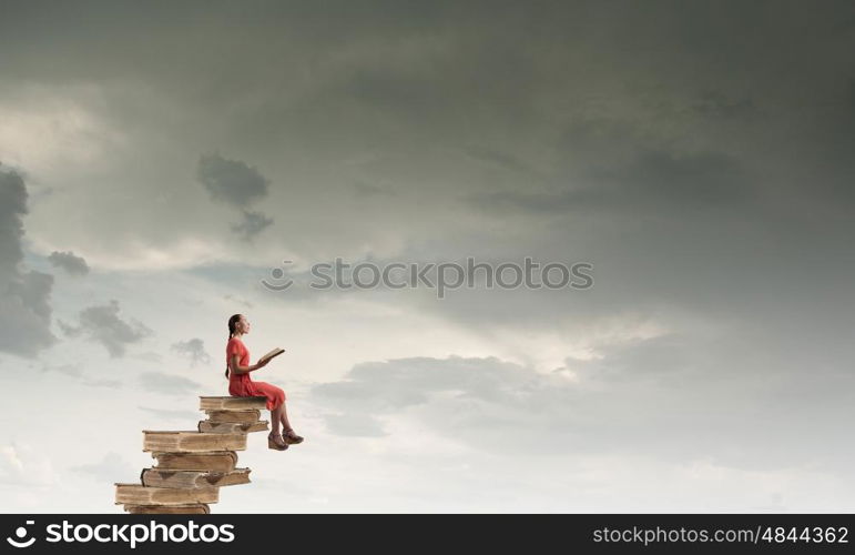 Attractive girl reading book. Young girl in red dress sitting on stack of books and reading