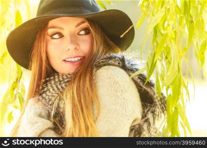 Attractive girl warm herself. Autumnal female fashion. Pretty young woman wearing stylish sweater waistcoat and hat warm herself outdoor. Beauty fashionable woman standing in park around leaves of willow tree.