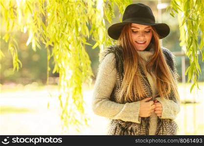 Attractive girl warm herself. Autumnal female fashion. Pretty young woman wearing stylish sweater waistcoat and hat warm herself outdoor. Beauty fashionable woman standing in park around leaves of willow tree.