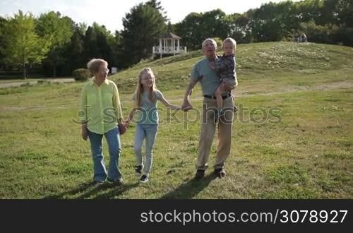 Attractive grandparents bonding with grandchildren while taking a walk in park. Joyful grandfather carrying his cute toddler grandson, holding hands of happy granddaughter while multi generation family walking in park and enjoying sunny day.