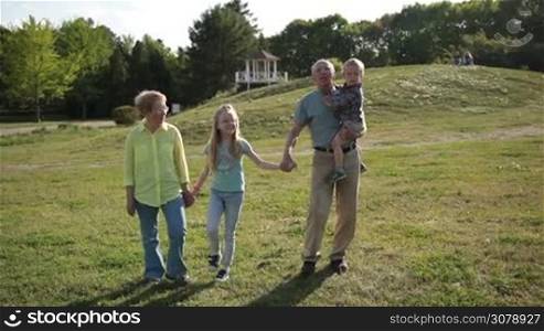 Attractive grandparents bonding with grandchildren while taking a walk in park. Joyful grandfather carrying his cute toddler grandson, holding hands of happy granddaughter while multi generation family walking in park and enjoying sunny day.