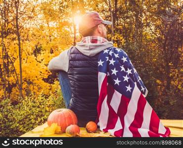 Attractive guy holding a US flag against the background of yellow trees and the setting sun. National holiday concept. Guy holding a US flag. National holiday