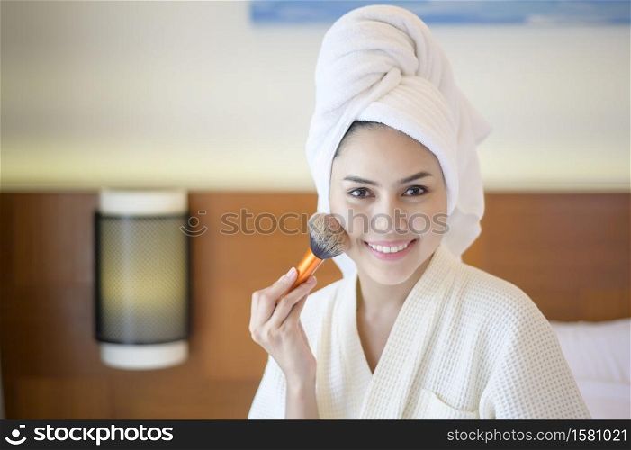 Attractive happy female in white bathrobe is applying natural Make-Up with cosmetic powder brush, Beauty Concept.