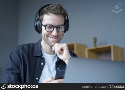 Attractive happy German home office worker in headphones and glasses sincerely smiling when looking at mobile phone screen in his hands while working remotely on laptop online. Freelance concept. Happy home office worker in headphones and glasses sincerely smiling when looking at phone screen