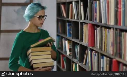 Attractive hipster college female student in stylish spectacles looking for books in library. Young woman with blue hair picking up books from the shelf in university library. Girl with pile of books in her hand choosing books from the shelves.