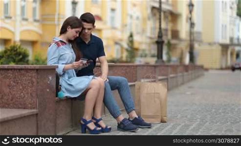 Attractive hipster couple making online purchase with mobile phone outdoors. Beautiful woman holding credit card and using smartphone for online shopping while spending leisure together with her handsome boyfriend.