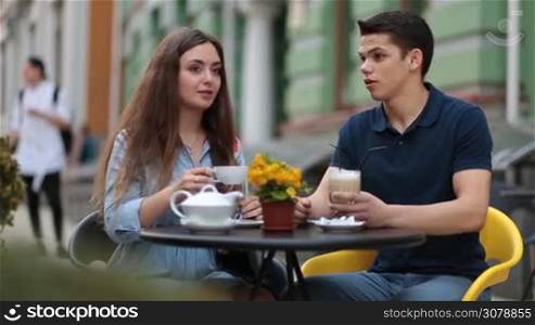 Attractive hipster couple sitting at the table in sidewalk cafe and talking. Romantic couple on travel during holidays. Affectionate spending leisure in coffee shop with mug of hot tea and coffee outdoors.
