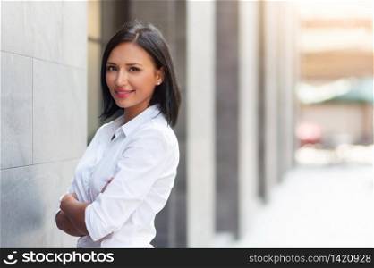 Attractive Hispanic female with arms crossed and looking at camera over building background at outdoor. Smiling Beautiful woman in white shirt standing with confident expression and copy space.