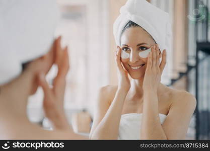 Attractive hispanic woman with eye patches looking at mirror during morning skincare routine. Smiling pretty female wrapped in towel after shower reducing facial wrinkles moisturizing skin under eyes.. Attractive latina woman with under eye patches looking at mirror during daily skincare treatment