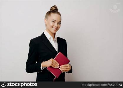 Attractive intelligent female project manager in dark suit with hair in bun and red notebook in hands standing in front of light wall and expressing happiness, smiling at camera while posing indoors. Happy business woman in dark suit posing indoors