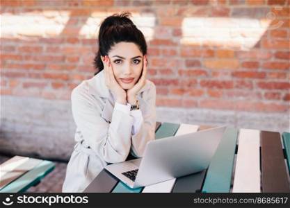 Attractive lady with dark hair and make-up, long red nails holding her hands on cheeks resting at cafe sitting at table in front of open laptop waiting for video call from her relatives or friends