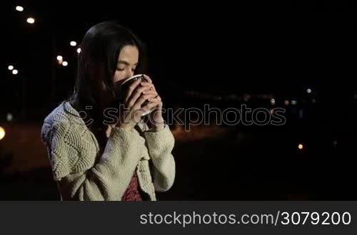 Attractive long brown hair girl drinking cup of fresh coffee while enjoying amazing urbanscape at night. Gorgeous young woman holding paper cup with hot drink and sipping during leisure on warm summer night outdoors. Slow motion. Stabilized shot.