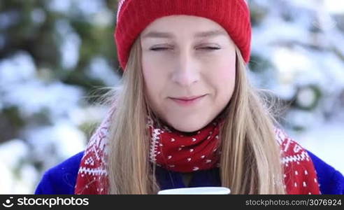 Attractive long hair blonde woman with beautiful deep blue eyes enjoying the scent of fresh hot coffee outdoors in the park in winter. Pretty girl in red scarf and hat holding and smelling a cup of coffee in knitted mittens in winter park.