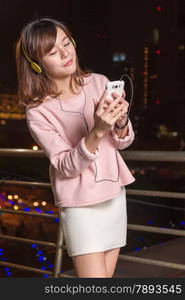 Attractive Malaysian female wearing headphones and holding a cell phone with city lights in background