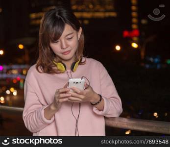 Attractive Malaysian female wearing headphones and holding a cell phone with city lights in background