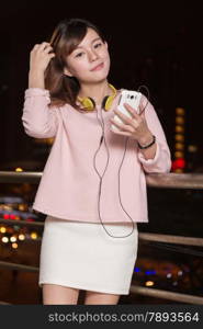 Attractive Malaysian female wearing headphones and holding a cell phone with city lights in background