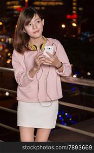 Attractive Malaysian female wearing headphones and holding a cell phone with city lights in background