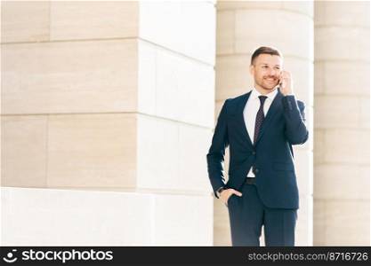 Attractive male CEO solves working issues with business partner during phone conversation, dressed in formal black suit, keeps hand in pocket, looks happily into diastance. Business and people