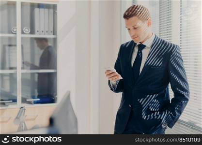 Attractive male director dressed in corporate clothing, checks email on mobile phone, stands near window with shutter in his cozy cabinet, blogs website, shares news with colleagues, uses internet